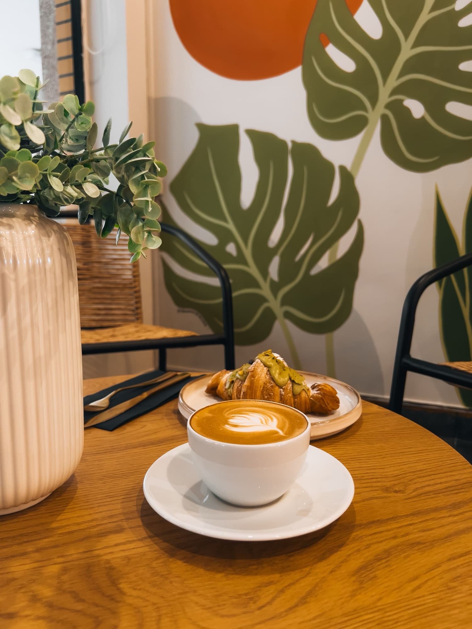 A cafe table with a latte and a pastry, warm wood and greenery behind
